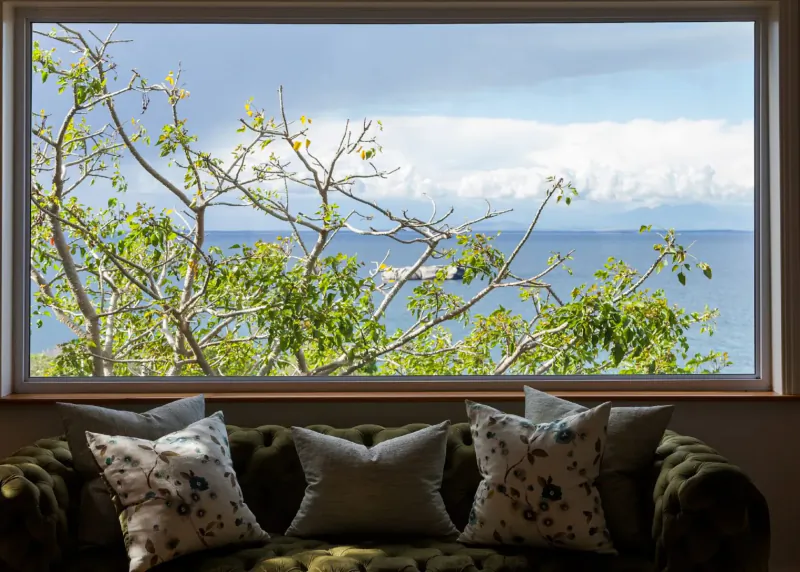 Window framing ocean view with scattered clouds and green tree branches, tufted green sofa with pillows in foreground at coastal retreat.