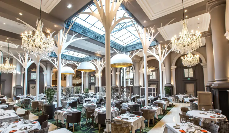 Elegant Saint-Malo gallery dining room with white tree columns, chandeliers, skylight, and round tables set for meals.