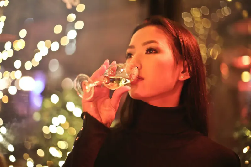 Asian woman with long red hair sips white wine from a glass, smiling in festive holiday lights setting
