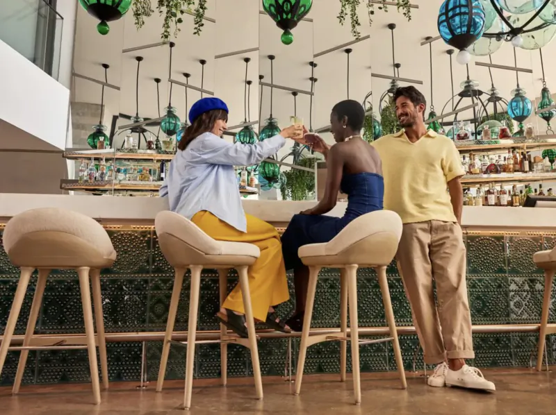 Three diverse people toasting drinks at a stylish bar with green lanterns and plants, W Algarve Hotel.