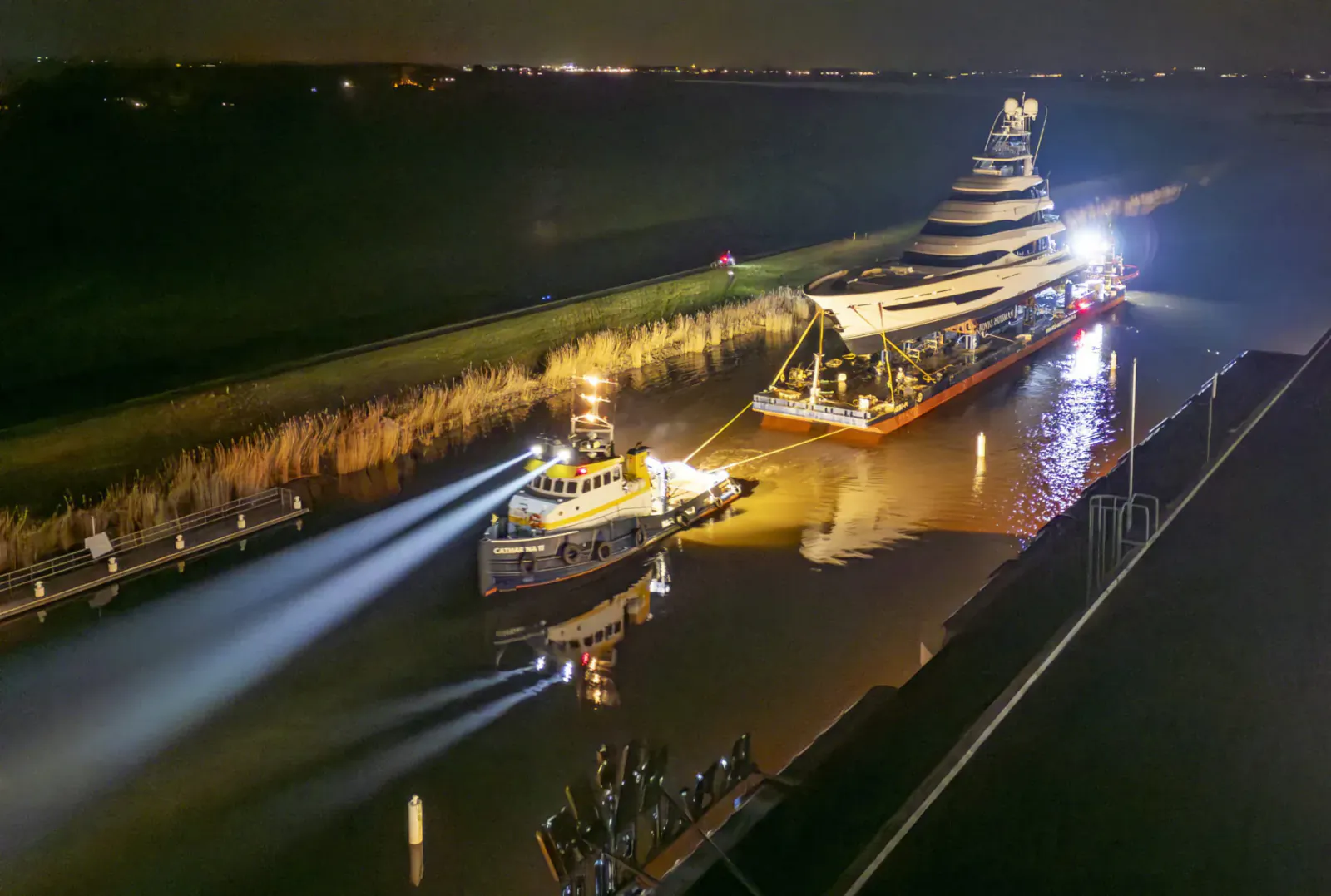 Night scene of illuminated multi-tiered barge towed by tugboat on canal, lights reflecting on water.