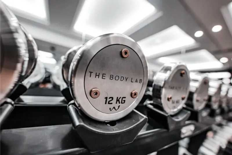 Close-up of The Body Lab 12 kg dumbbells on gym rack, branded 'The Body Lab 12 KG'.