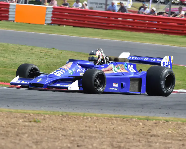 Blue 1970s Formula 1 race car with 'B.A.R.' logo speeding on track at motorsport event, surrounded by barriers and spectators.