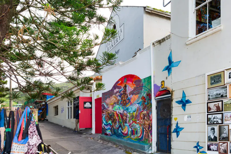 Olympia Café gallery in Kalk Bay: vibrant mural on alley wall with pine trees, blue origami, photos, and hanging clothes