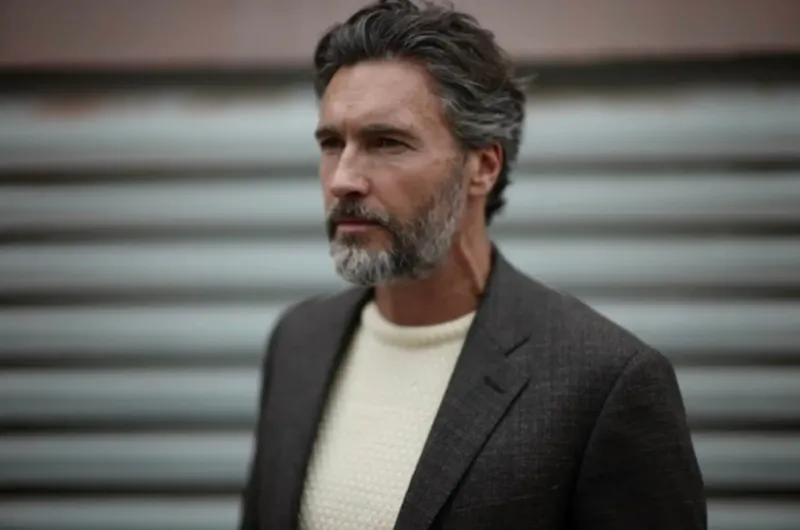 Man with salt-and-pepper beard and hair in grey check blazer over cream sweater, looking aside against corrugated wall