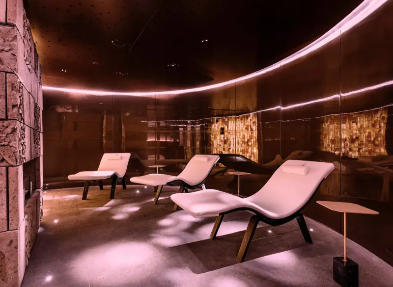 Modern spa lounge with three white reclining chairs, pink ambient lighting, mirrored walls, and salt stone accents.