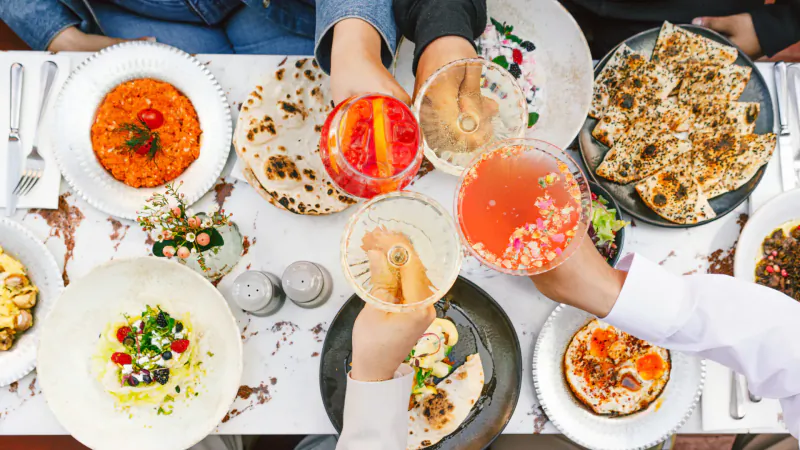 Overhead view of hands clinking colorful cocktails amid Middle Eastern dishes like naan, curries on white table.