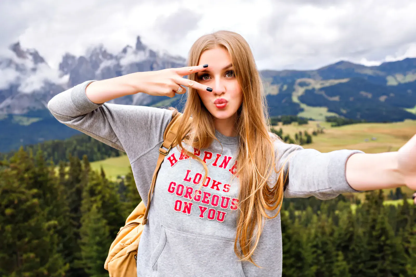 Blonde woman in 'Happine Looks Gorgeous on You' sweatshirt making peace sign and duck face in Alps mountains