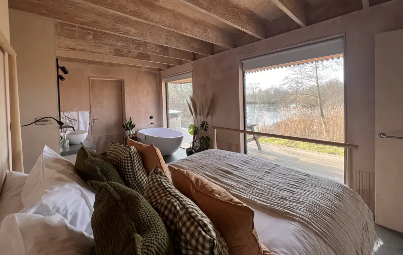 Cozy wooden glamping bedroom with king bed, freestanding bathtub, and lake view through large windows.