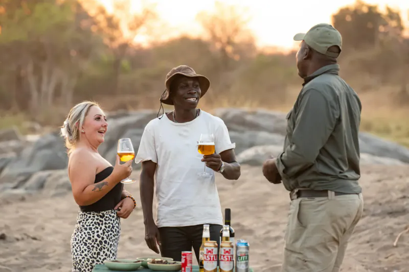 Three smiling people—a white woman, Black man in hat, Black man in cap—toasting drinks at sunset on rocky hill with beers and bottles.