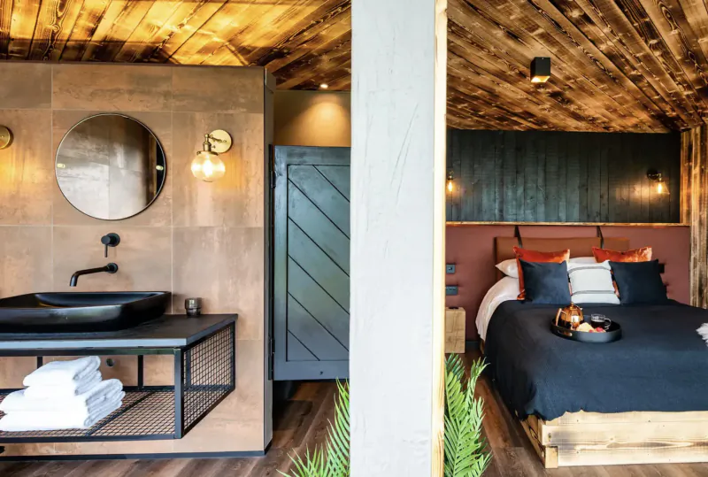 Split-view of The Boathouse luxury lodge: wooden bathroom with black sink, mirror, towels; adjacent bedroom with navy bed, orange pillows.