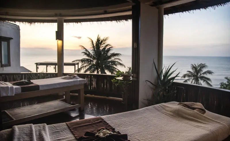 Two massage tables on open-air balcony overlooking sunset ocean view with palm trees