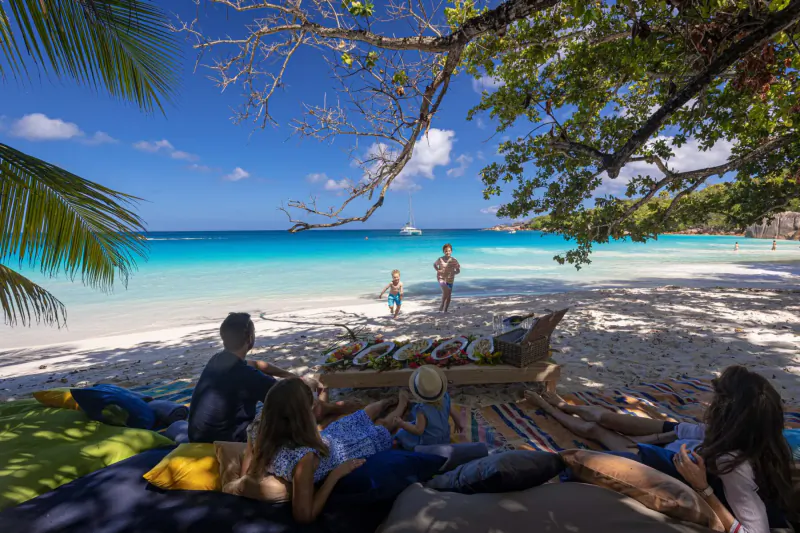 Family picnicking on white sand beach under palm trees at turquoise Praslin lagoon, Seychelles, with kids playing in water.