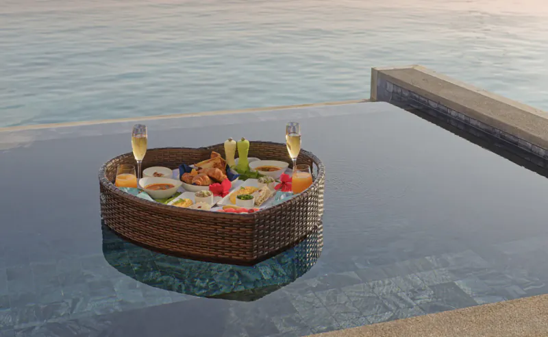 Heart-shaped woven basket with champagne flutes, veggie crudités, and snacks floating in infinity pool overlooking ocean at Amari Raaya Maldives