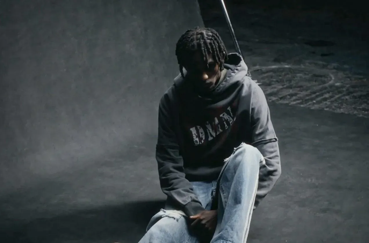 Man with dreads sits pensively in BALENCIAGA hoodie and jeans, dark studio setting for DEMN STUDIOS launch