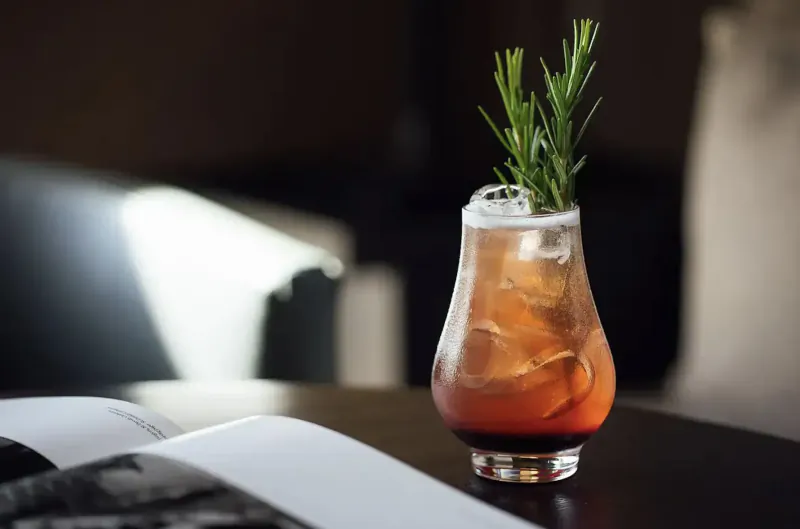 Amber cocktail garnished with rosemary sprig in elegant glass on wooden table with open book, beside sofa in art hotel gallery.
