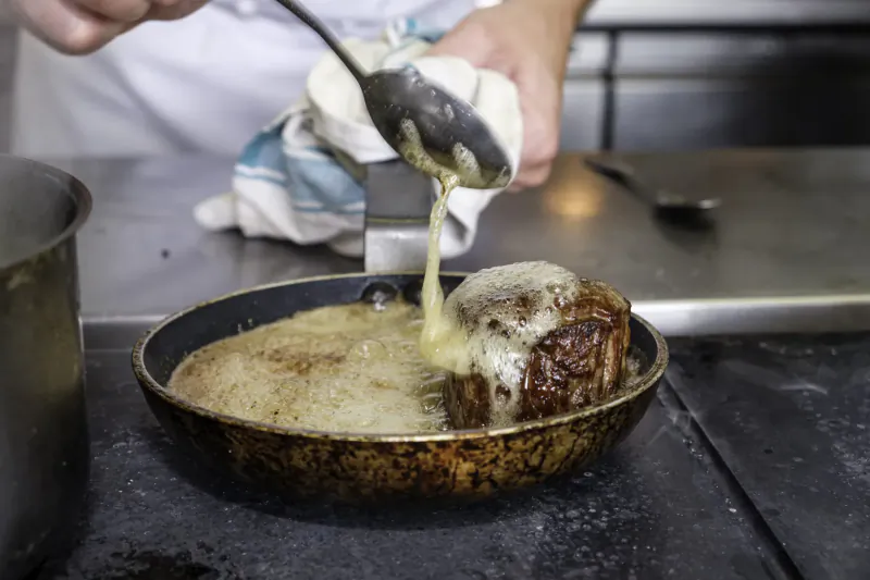 Chef pouring thick sauce from ladle over seared steak with melted cheese in black pan on kitchen counter