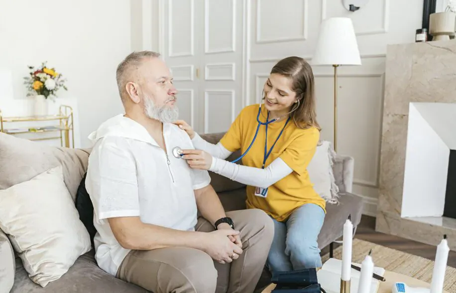 Nurse in yellow uniform listening to heartbeat of smiling senior man seated on couch in elegant living room
