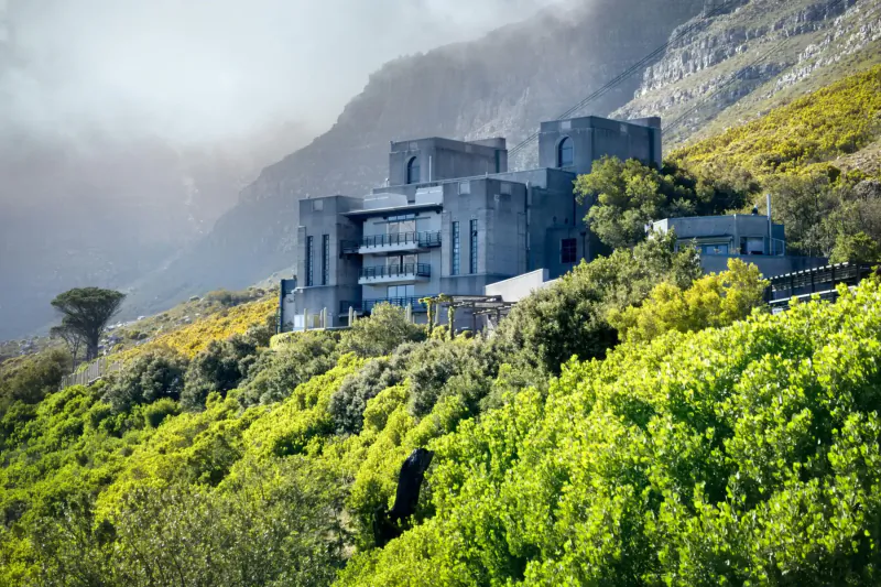Modern castle-like building perched on lush green mountainside near Cape Town, with misty peaks and fynbos vegetation.
