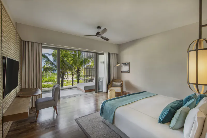 Spacious Anantara Iko Mauritius resort bedroom with teal bed, chairs, TV, and sliding doors to beach view.