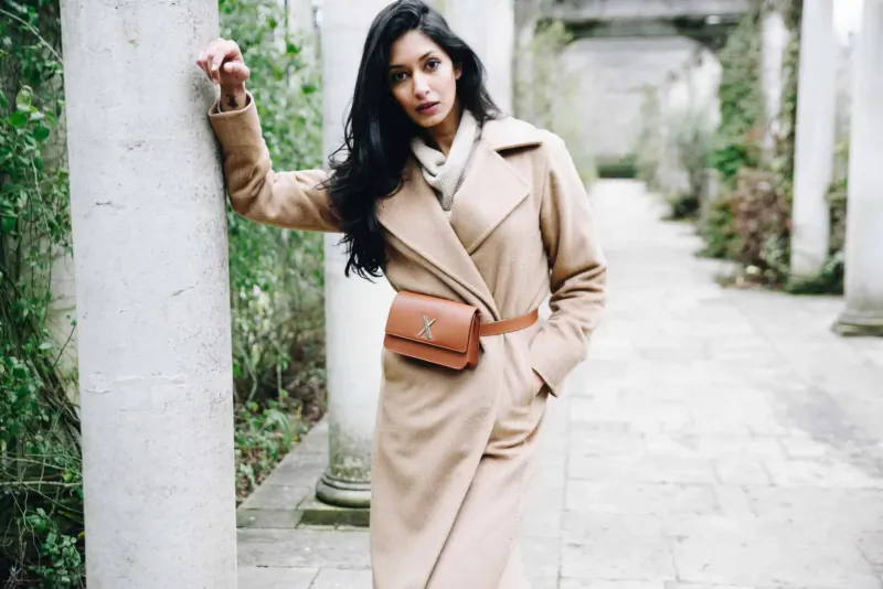 Woman in beige coat and tan belt bag leans against pillar in leafy colonnade, Luxtra sustainable fashion