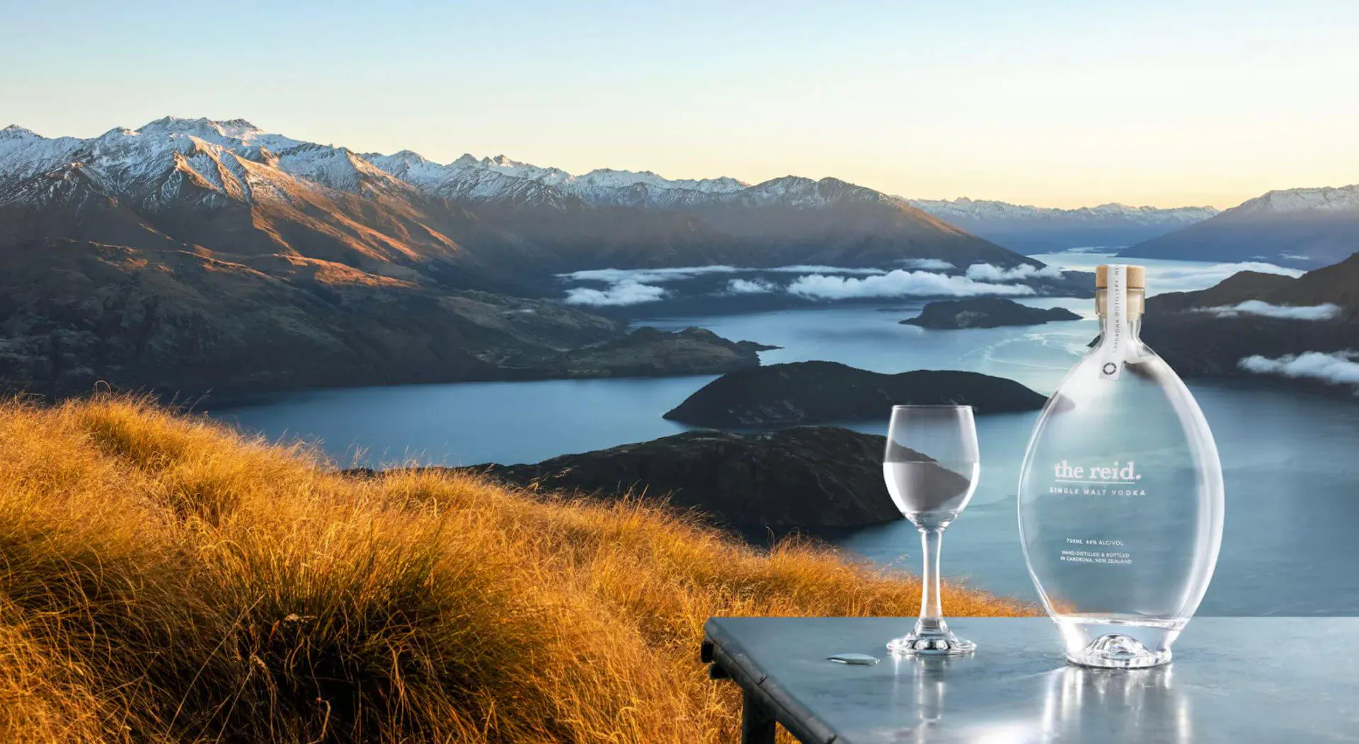 Cardrona Distillery vodka bottle and glass on table overlooking snowy mountains, lake, golden grasses at sunset, New Zealand.