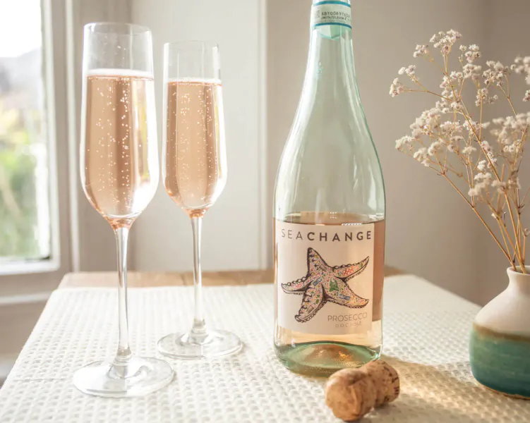 Two glasses of fizzy rosé champagne beside Sea Change rosé wine bottle with starfish label, cork, and white flowers on table.