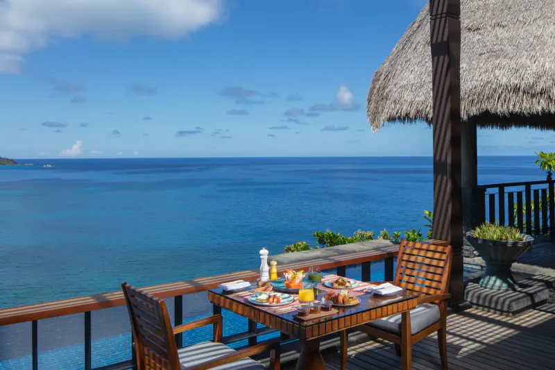 Oceanfront breakfast table on Anantara Seychelles resort balcony with tropical fruits, pastries, and blue sea view.
