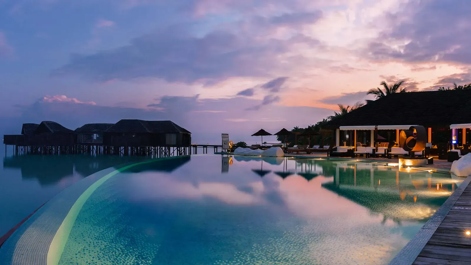 Twilight infinity pool at Lily Beach Resort & Spa reflecting overwater bungalows, palm trees, and sunset sky.