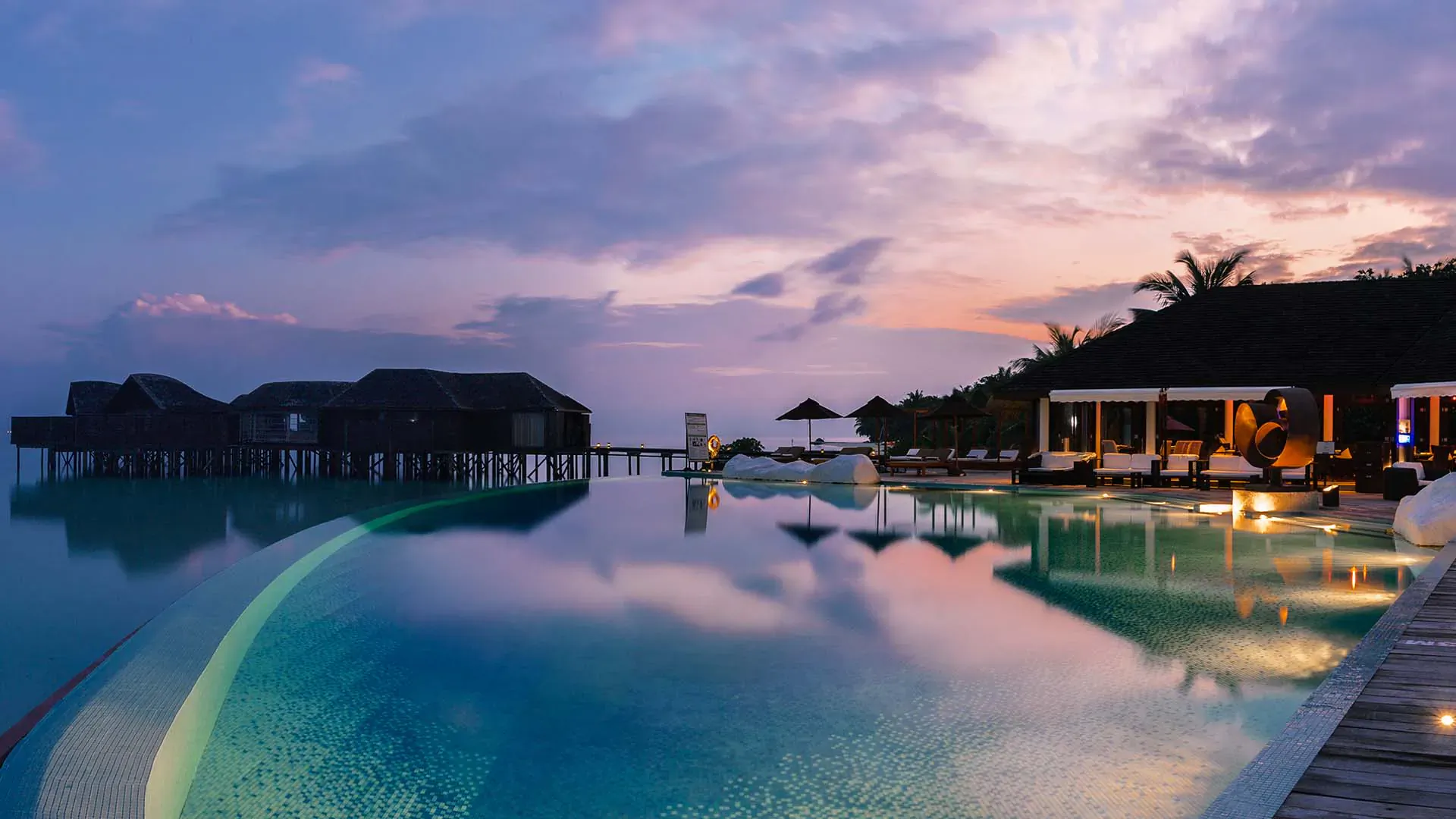 Twilight infinity pool at Lily Beach Resort & Spa reflecting overwater bungalows, palm trees, and sunset sky.