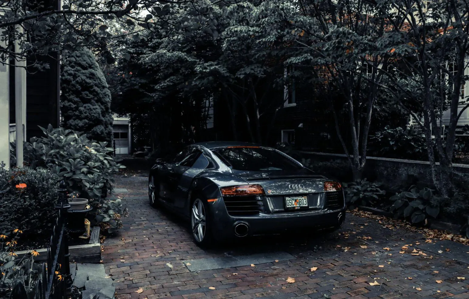 Black Audi R8 sports car parked on brick driveway in lush green garden courtyard, autumn leaves scattered.