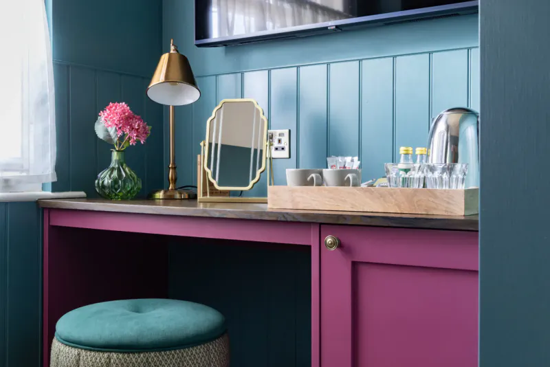 Luxury hotel vanity with pink desk, gold mirror, lamp, pink roses, teal walls, and tea set.