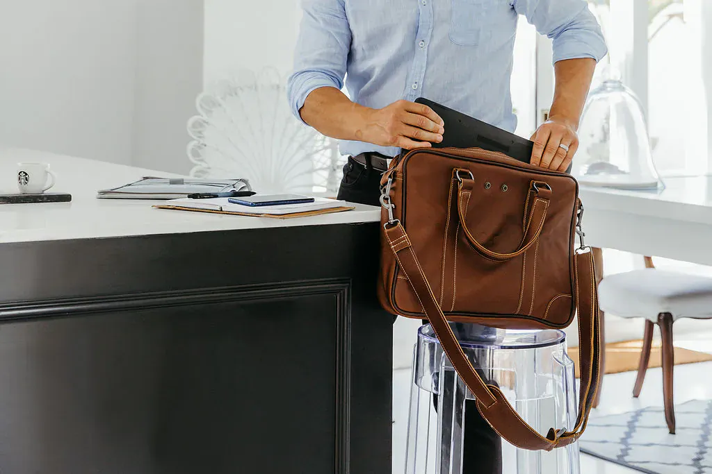 Man in light blue shirt placing tablet into brown Thandana leather briefcase at modern office desk