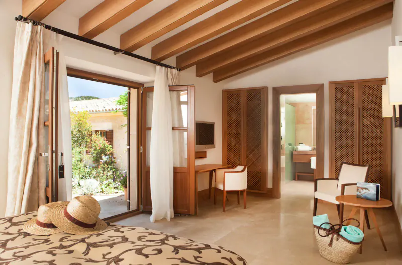 Luxury Mallorca hotel room with wooden beams, open doors to garden, bed with straw hat, desk, and chairs.