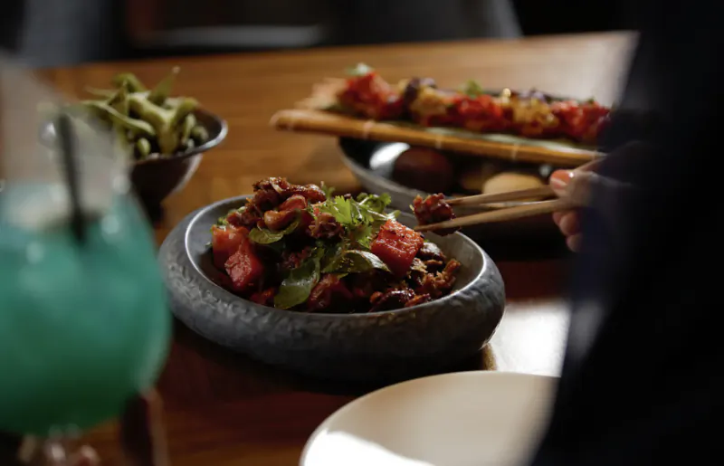 Close-up of Myazú Restaurant table: edamame, spicy skewers on bamboo, kidney beans with greens in stone bowl, chopsticks, blue drink.