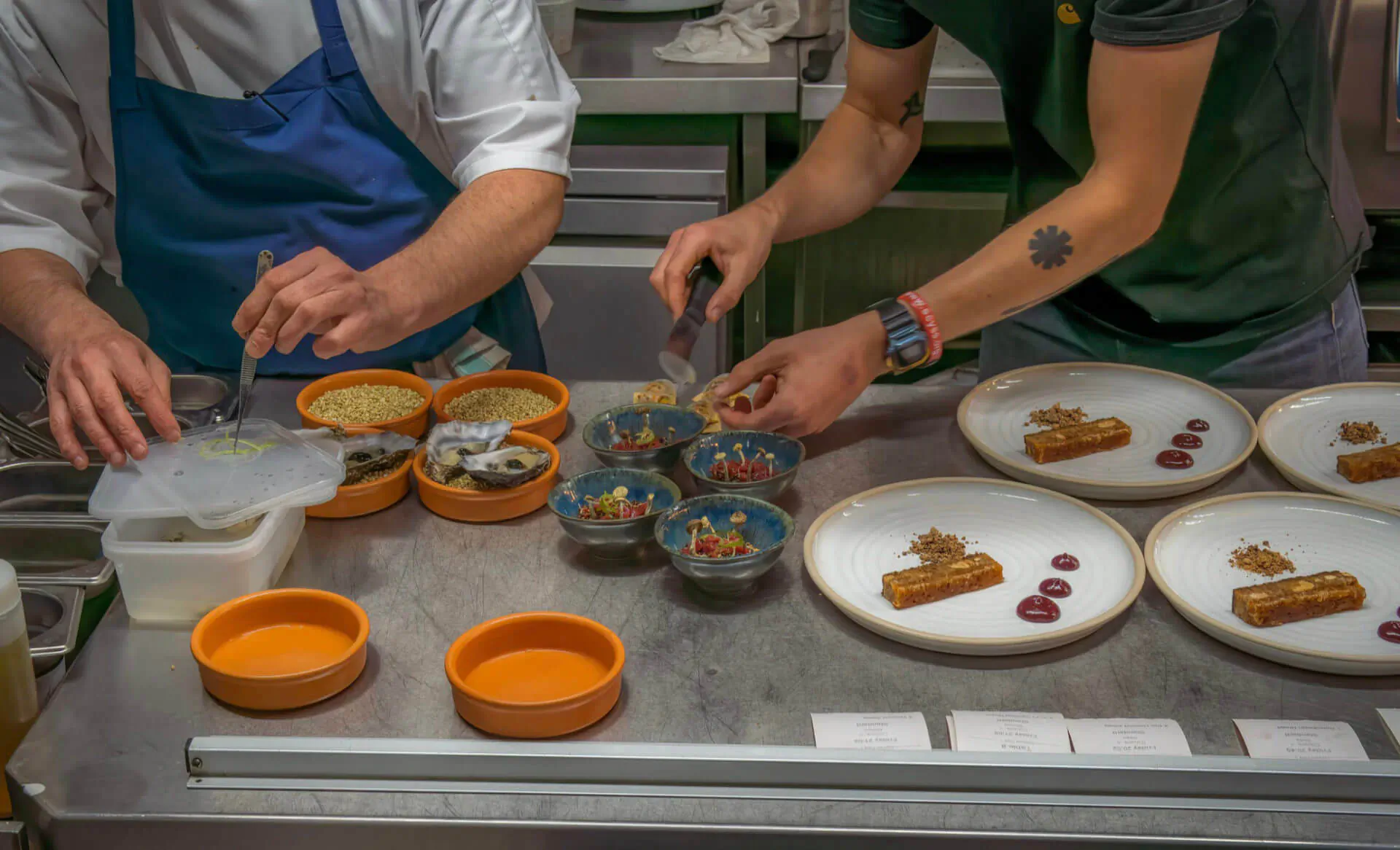 Two chefs in white uniforms and aprons prepare plated dishes with orange bowls and garnishes on stainless steel counter at 1771 Restaurant.