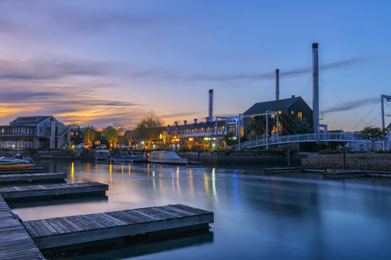 Twilight view of Turbine Hotel: brick buildings with smokestacks, white bridge over calm marina with docked boats and wooden docks.