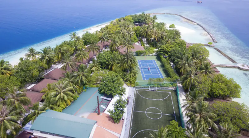 Aerial view of Lily Beach Resort & Spa: blue tennis court, green football pitch, thatched villas, palm trees, turquoise lagoon.