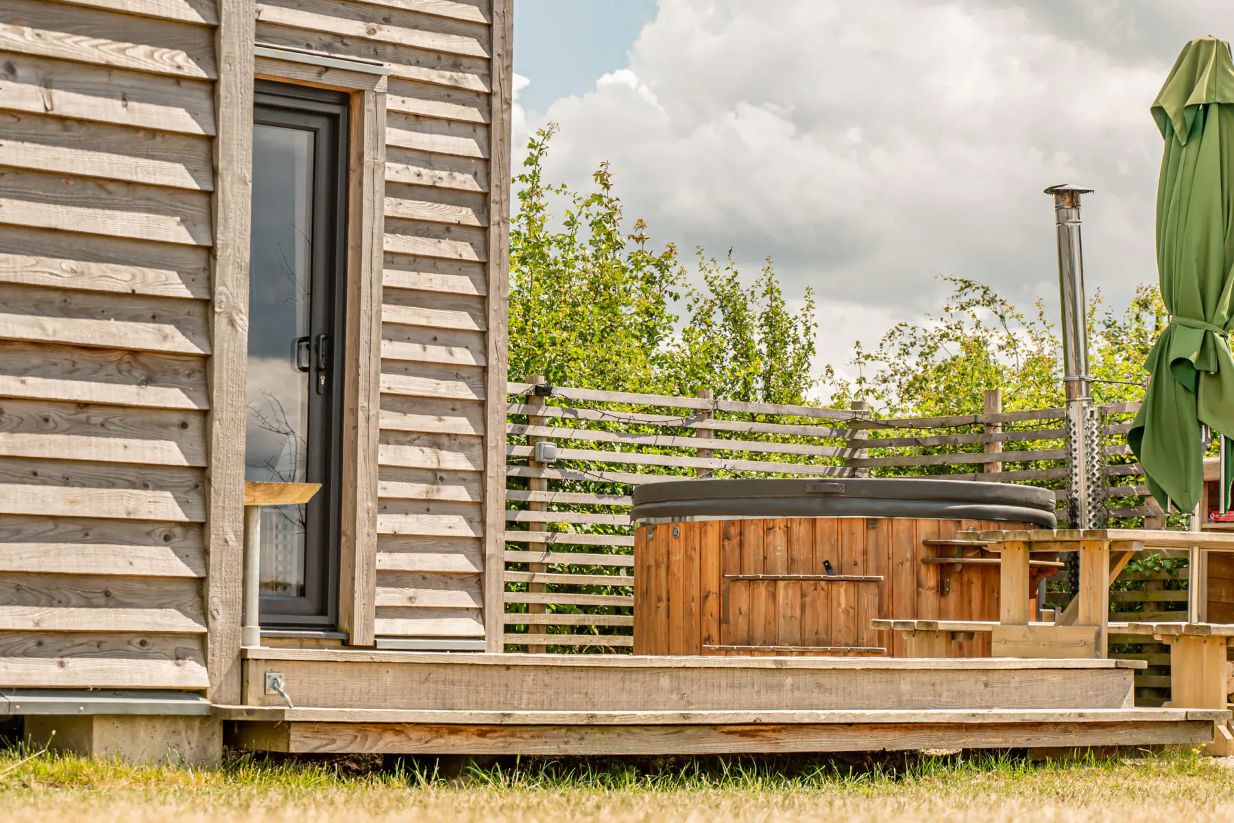 Rustic wooden cabin with hot tub, green umbrella, picnic table on deck amid grassy field and trees under cloudy sky