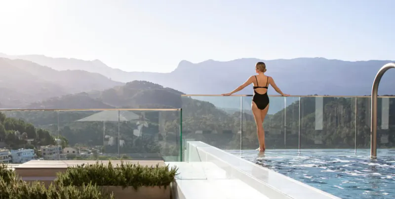 Woman in black bikini stands at glass railing of rooftop infinity pool, overlooking Mallorca mountains at Jumeirah Port Soller Hotel & Spa