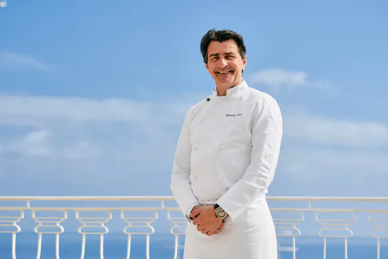 Smiling chef in white uniform with 'Moulin' text, hands clasped, standing by balcony railing under blue sky in Monaco