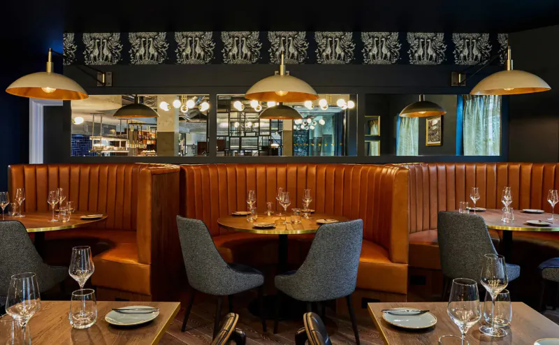 Elegant Hotel Indigo Chester restaurant with orange tufted booths, round tables set with wine glasses, brass pendant lights, and open kitchen view.