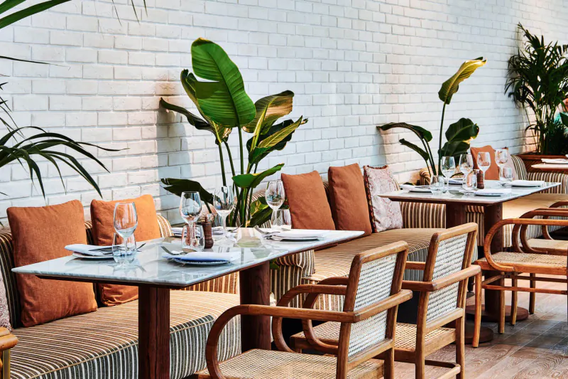 Elegant outdoor dining area at 25hours Hotel Dubai with wicker chairs, glass tables set with wine glasses, orange cushions, and lush banana plants against white brick wall.