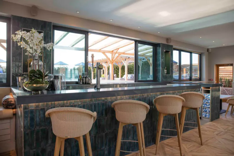Elegant indoor bar at Manor House Hotel with teal counter, cream stools, orchids, and pool view through glass doors.