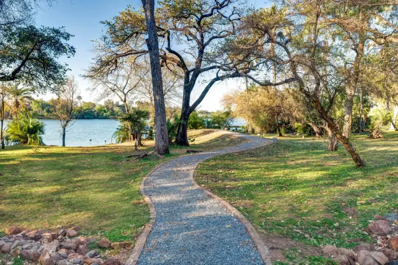 Curving gravel path through grassy park with trees and lake near Victoria Falls at Palm River Hotel