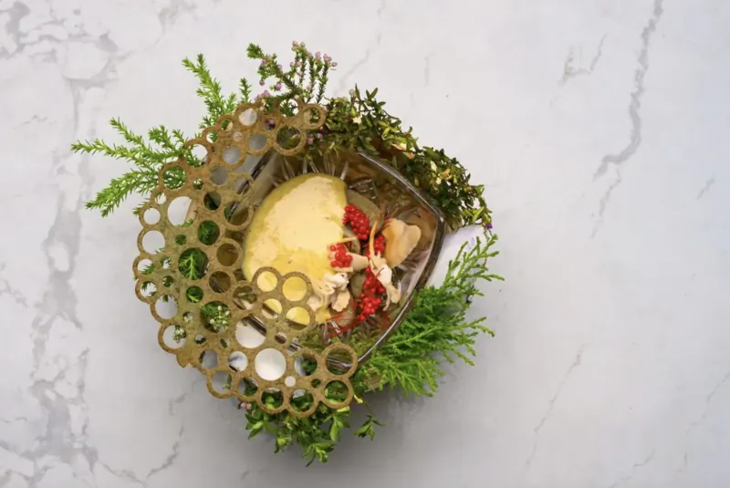 Foraged dish with sliced yellow fruit, red berries, and ferns in ornate gold perforated bowl on marble surface