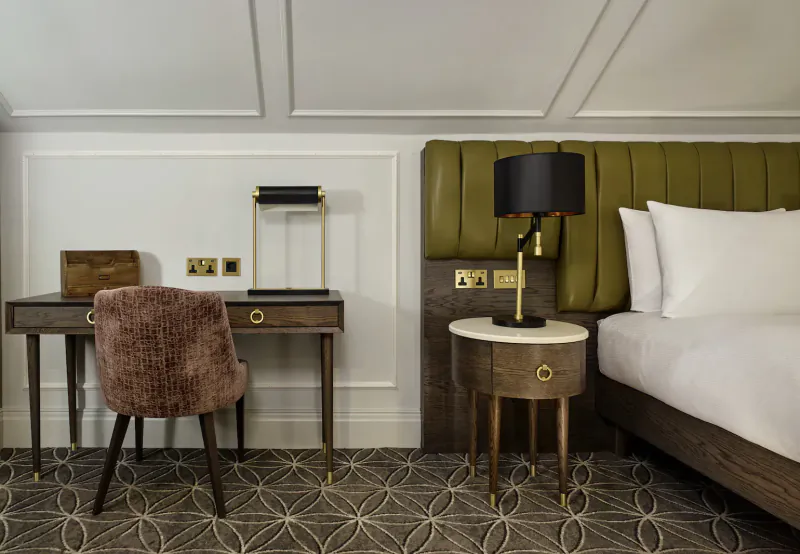 Elegant hotel room with wooden desk, plush chair, green upholstered bed, and brass lamps.