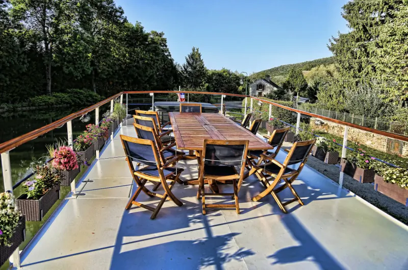 Wooden dining table with chairs on sunny river cruise deck balcony, surrounded by flowers, trees, and Burgundy valley views.