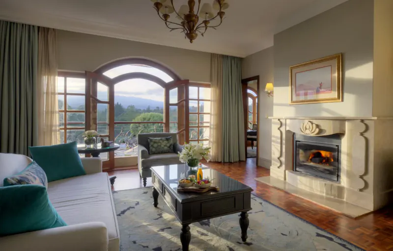 Elegant hotel lounge with white sofa, coffee table with snacks, lit fireplace, chandelier, and mountain view through arched windows.