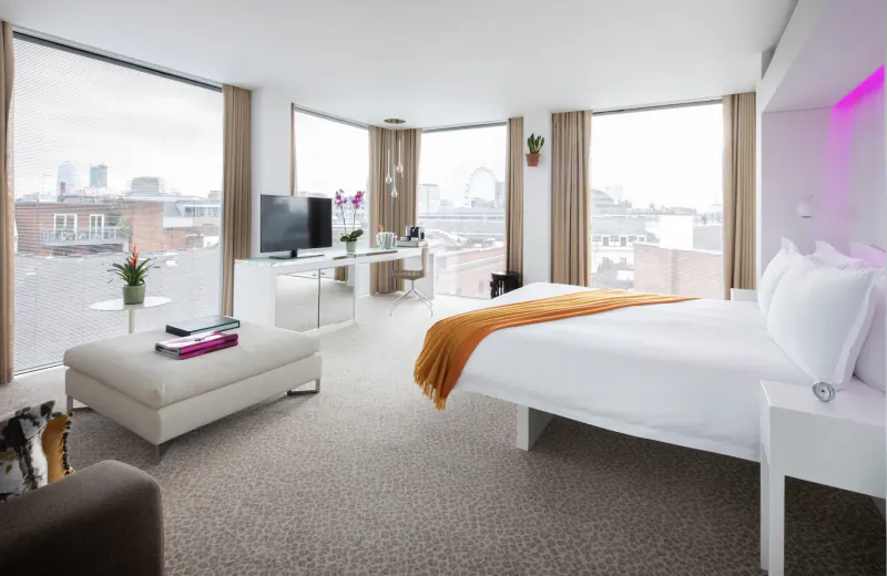 Modern luxury hotel room with panoramic London skyline view, white bed with orange throw, neon pink accents, orchids, and TV.